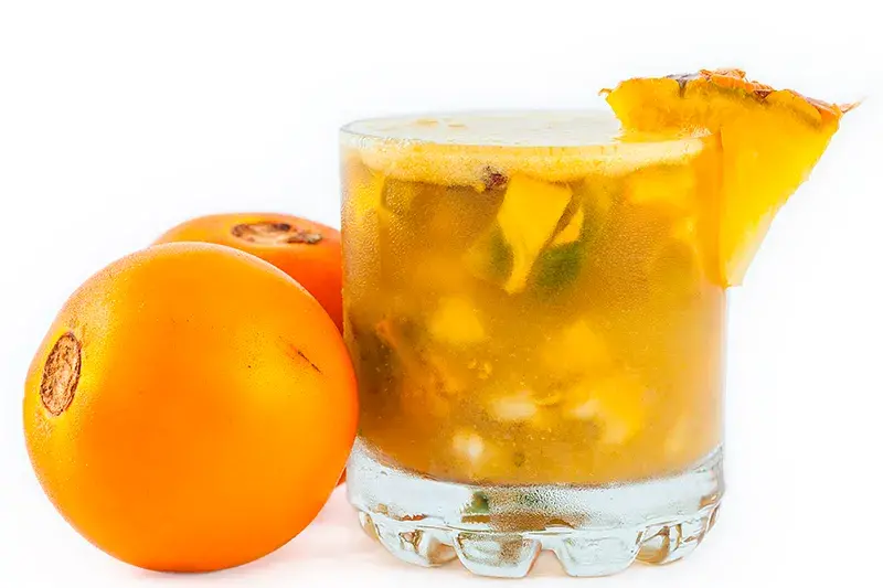 Vaso de champús con trozos de frutas y dos lulos enteros al lado, decorado con piña.