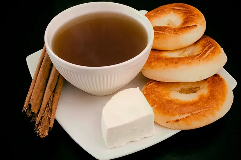 Taza de aguapanela caliente con almojábanas, queso y ramas de canela en un plato blanco.