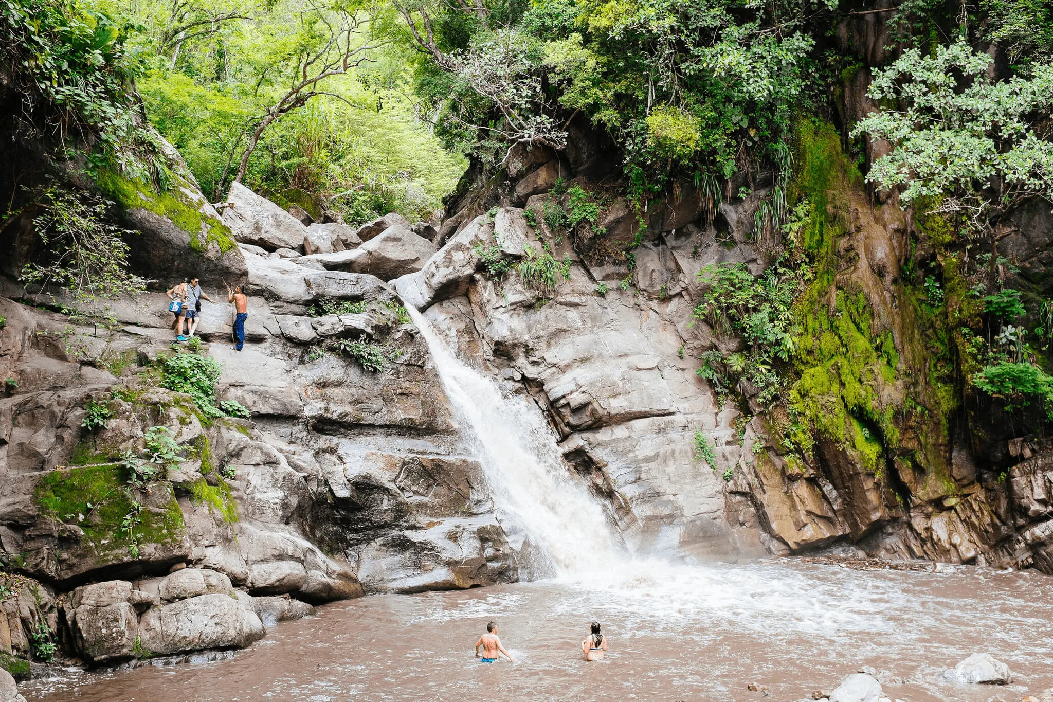 Cascada en la selva con personas bañándose en la poza y otras en las rocas.