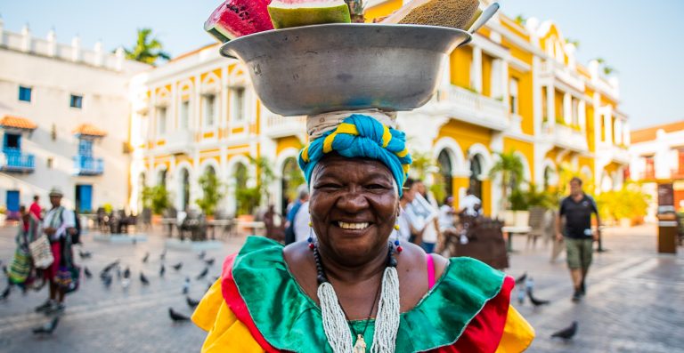 fotografía de palenquera sonriendo con frutas sobre su cabeza con fondo de plaza de bolivar en Cartagena, día de la raza