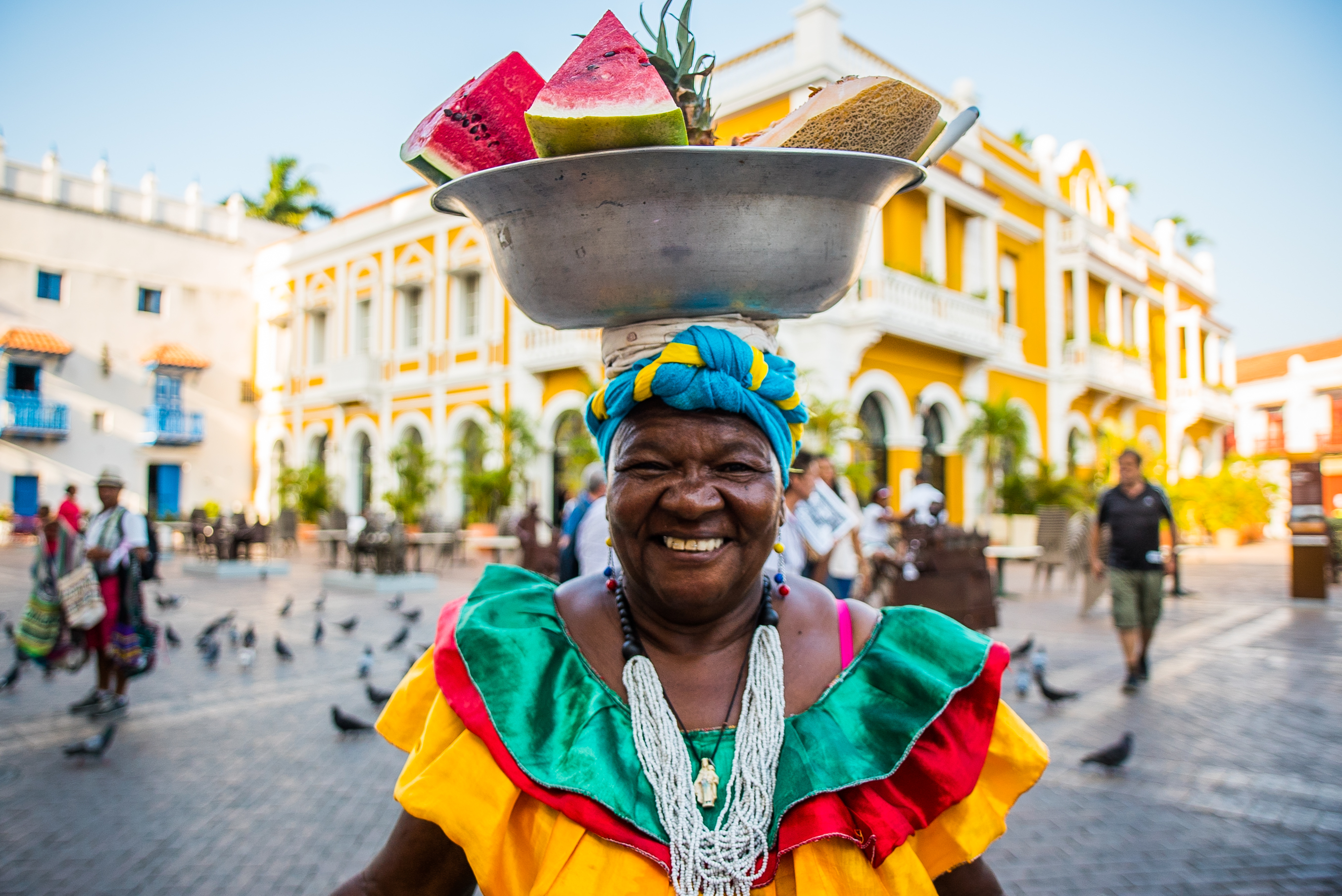 fotografía de palenquera sonriendo con frutas sobre su cabeza con fondo de plaza de bolivar en Cartagena, día de la raza