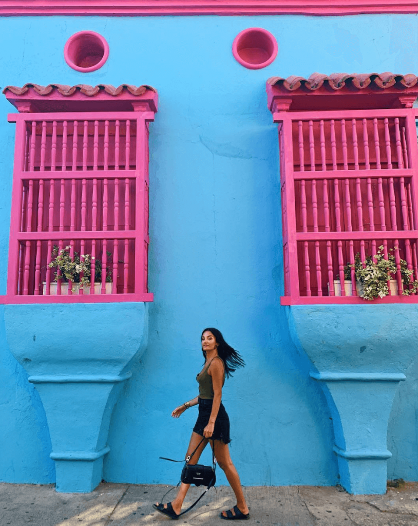 Amina Muaddi fashionista Europea en Colombia paseando frente a una casa azul con ventanas rosadas, Cartagena, moda Colombia