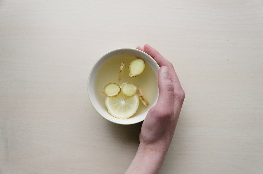 mesa blanca con taza de té de Jengibre que contiene pedazos de este encima y una mano sosteniendola, jengibre, remedios caseros