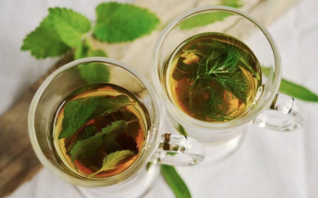 fotografía de té de hierbas en mesa con hojas de menta adentro sobre mesa blanca que tiene hojas regadas , remedio casero
